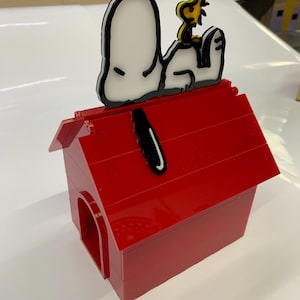 May include: A red doghouse made of interlocking blocks, with a Snoopy and Woodstock figure on top. The Snoopy figure is white with black outlines, and Woodstock is yellow. The doghouse has a red roof and a rounded doorway.