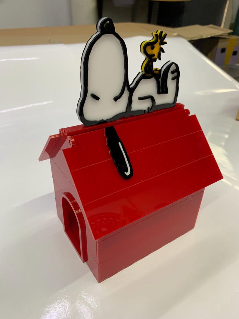 May include: A red doghouse made of interlocking blocks, with a Snoopy and Woodstock figure on top. The Snoopy figure is white with black outlines, and Woodstock is yellow. The doghouse has a red roof and a rounded doorway.