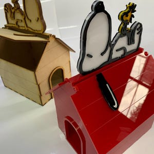 May include: Two dog houses, one in natural wood and the other in red, each with a Snoopy and Woodstock design on top. The red dog house has a glossy finish. The Snoopy and Woodstock figures are black, white, and yellow.