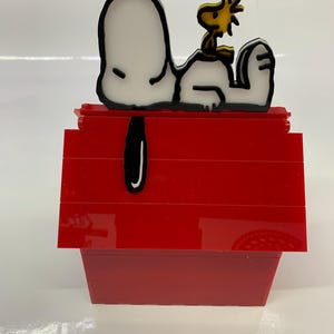 May include: A red doghouse with a Snoopy and Woodstock design. Snoopy is white with a black outline, and Woodstock is yellow with a black outline. The doghouse is made of red blocks.