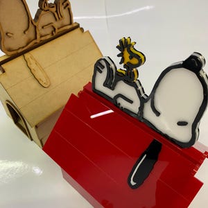 May include: Two Snoopy dog houses. One is made of light brown wood, and the other is red. Both have Snoopy and Woodstock figures on top. The Snoopy figure is white with black accents, and Woodstock is yellow.