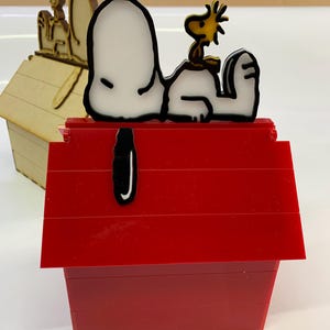 May include: A red doghouse-shaped box with a black and white Snoopy figure and a yellow Woodstock figure on top. A second, smaller doghouse is visible in the background, made of light brown wood.