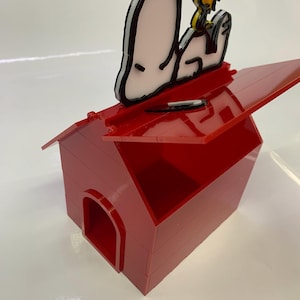May include: A red acrylic dog house with a hinged roof, featuring Snoopy and Woodstock figures on top. The figures are outlined in black and white, with yellow accents. The dog house has an arched doorway.