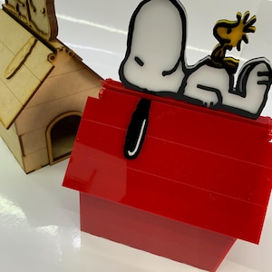 May include: A red doghouse with a white and black Snoopy figure and a yellow Woodstock figure on top. A second, wooden doghouse is in the background. The figures are likely decorative items.