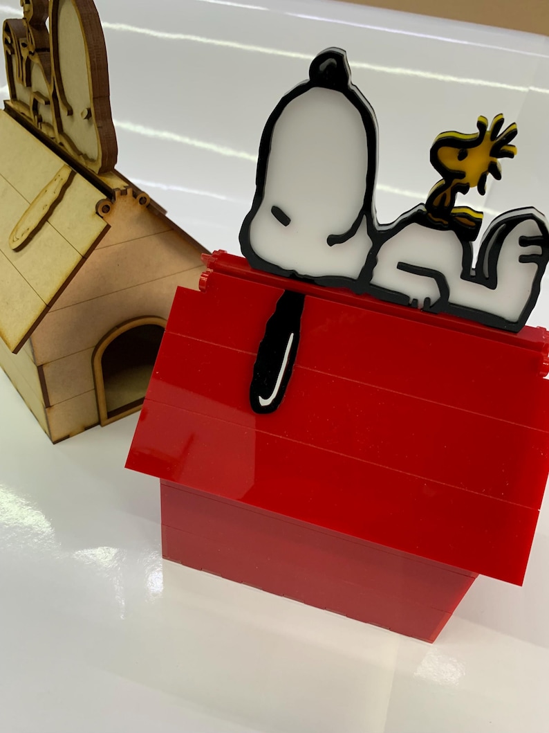 May include: A red doghouse with a white and black Snoopy figure and a yellow Woodstock figure on top. A second, wooden doghouse is in the background. The figures are likely decorative items.