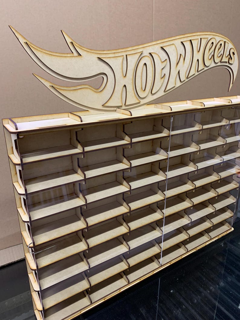 May include: A wooden Hot Wheels display shelf with clear acrylic panels. The shelf has 24 slots for storing Hot Wheels cars. The shelf is topped with a wooden Hot Wheels logo.