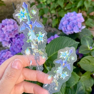 May include: Two clear stickers featuring a stylized sword design with blue and white floral accents. The sword has a blue hilt and wings, with delicate flowers and vines. The stickers are held in front of a backdrop of purple flowers and green foliage.