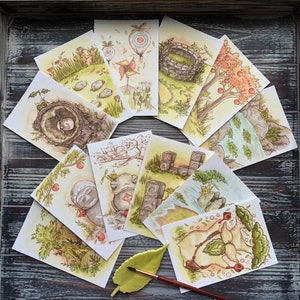 Op de afbeelding: Een verzameling aquarelprints met grillige sc&egrave;nes. De illustraties tonen landschappen met bomen, rotsen en fantastische elementen. Het kleurenpalet omvat groen, bruin en geel, met een zachte, dromerige esthetiek. De prints zijn in een waaier op een houten oppervlak gerangschikt.