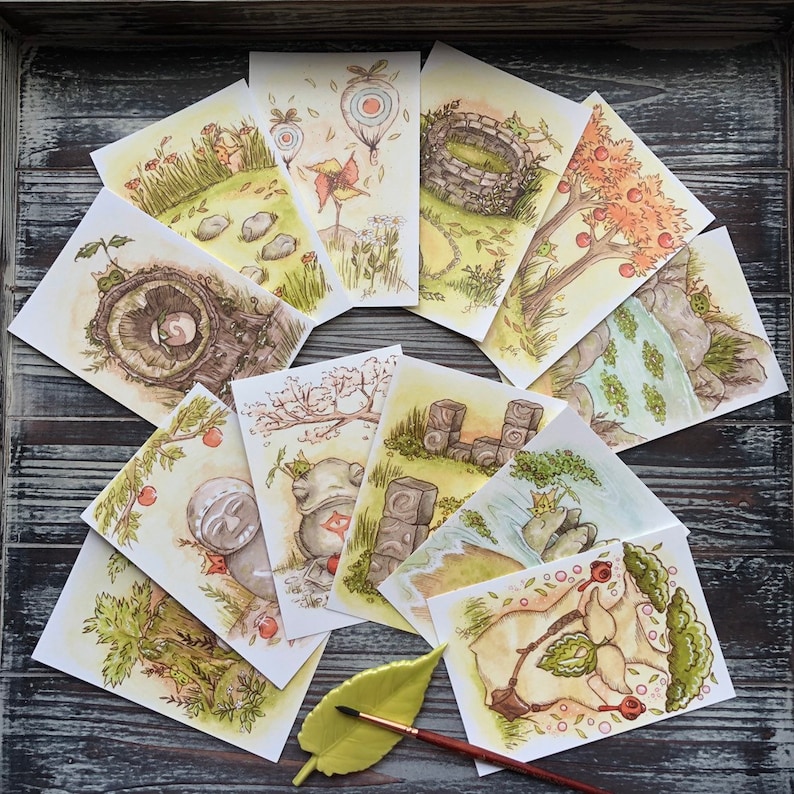 Op de afbeelding: Een verzameling aquarelprints met grillige sc&egrave;nes. De illustraties tonen landschappen met bomen, rotsen en fantastische elementen. Het kleurenpalet omvat groen, bruin en geel, met een zachte, dromerige esthetiek. De prints zijn in een waaier op een houten oppervlak gerangschikt.