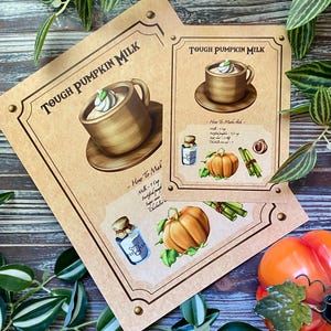 Op de afbeelding: Twee bruine papieren prints met de tekst "TOUGH PUMPKIN MILK" en illustraties van een kopje koffie, pompoen en ingredi&euml;nten. De prints zijn versierd met een bruine rand en messing klinknagels. Groene en witte bladeren omringen de prints.