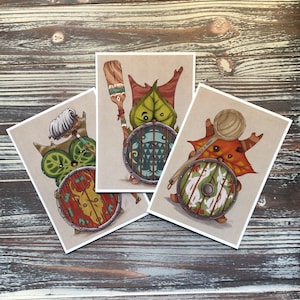 Puede incluir: Tres tarjetas ilustradas con criaturas fant&aacute;sticas con escudos. La primera tarjeta presenta una criatura verde con cabeza en forma de hoja sosteniendo un escudo con un dise&ntilde;o verde, azul y blanco. La segunda tarjeta presenta una criatura marr&oacute;n con un escudo con un dise&ntilde;o rojo, blanco y negro. La tercera tarjeta presenta una criatura marr&oacute;n con un escudo con un dise&ntilde;o blanco, negro y marr&oacute;n.