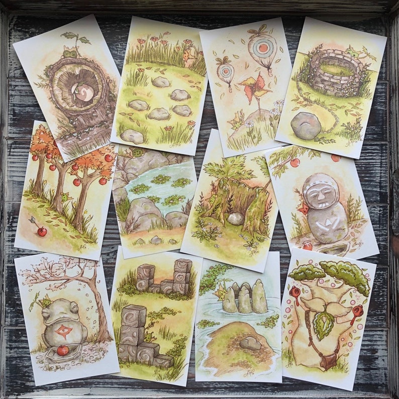 Op de afbeelding: Een verzameling aquarelkaarten met grillige natuurtaferelen. Elke kaart toont unieke illustraties van bomen, rotsen en wezens in een zacht, aards kleurenpalet. De kaarten zijn gerangschikt in een houten dienblad, wat een rustieke uitstraling cre&euml;ert.