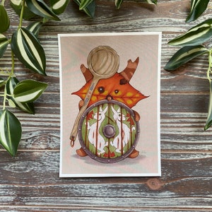 Puede incluir: Una ilustraci&oacute;n caprichosa de una criatura del bosque con un escudo y una cuchara de madera. La criatura se representa como una hoja marr&oacute;n y naranja con una cara y cuernos. El escudo est&aacute; decorado con dos conejos blancos y acentos rojos y verdes.