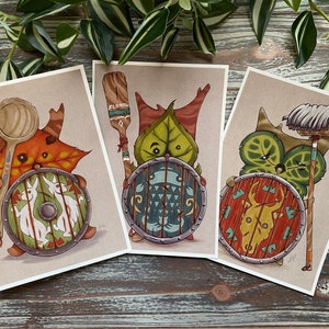 May include: Three illustrated prints featuring whimsical creatures with shields and weapons. The first creature is a brown and orange leaf creature holding a wooden spoon. The second creature is a green leaf creature holding a wooden paddle. The third creature is a green leaf creature holding a white mop.