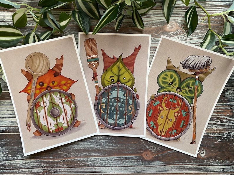 Puede incluir: Tres impresiones ilustradas con criaturas caprichosas con escudos y armas. La primera criatura es una criatura de hoja marr&oacute;n y naranja que sostiene una cuchara de madera. La segunda criatura es una criatura de hoja verde que sostiene una pala de madera. La tercera criatura es una criatura de hoja verde que sostiene una fregona blanca.