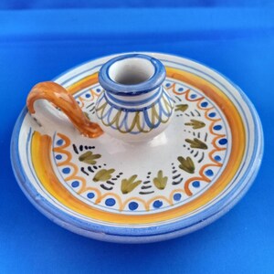 May include: A ceramic candle holder with a circular base and a central cup. The holder features a white background with orange, blue, and green painted details. It has a small handle and is set against a blue background.