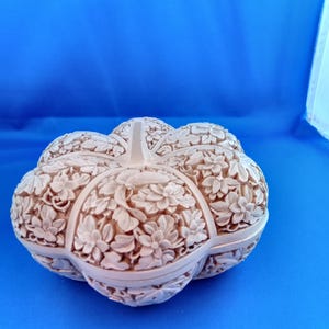 May include: A beige, intricately carved pumpkin-shaped box with floral patterns. The box has a stem and is set against a blue background. The detailed carvings create a textured appearance.
