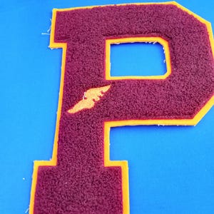 May include: A maroon chenille letter P patch with a yellow felt border. The letter features a textured, fuzzy surface and a small, yellow, wing-shaped accent detail on the left side of the vertical bar, set against a solid blue background.