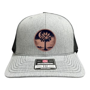May include: A gray and black trucker hat with a brown leather patch featuring a palm tree and crescent moon. The patch has the text "EST. 1970 RICHARDSON 112 ORIGINAL TRUCKER ADJUSTABLE".