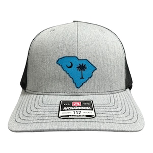 May include: Gray and black trucker hat with a blue South Carolina state outline and a palm tree. The hat has a black mesh back and a black adjustable strap with a Richardson 112 logo.