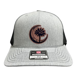May include: A gray and black trucker hat with a brown patch featuring a palmetto tree and crescent moon. The hat has a black mesh back and a black adjustable strap with a Richardson 112 label.