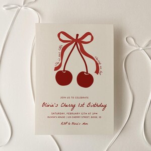 Puede incluir: Una tarjeta de invitación blanca con ilustraciones de cerezas rojas y un lazo de cinta rojo. El texto en la tarjeta dice "Únete a nosotros para celebrar el primer cumpleaños de Olivia's Cherry, Sábado 12 de febrero a las 3 PM, Olivia's House 129 Cherry Street Boise, ID, RSVP a Olivia's Mom."