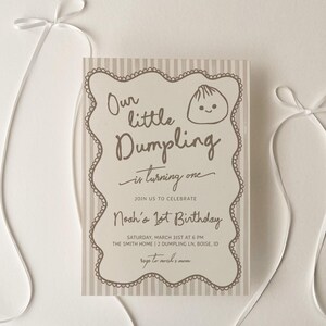May include: A light-colored invitation card with the text "Our little Dumpling is turning one" and details for a first birthday celebration. The card is decorated with a scalloped border and a dumpling illustration. White ribbons are arranged around the card.