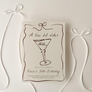 Puede incluir: Una tarjeta de invitación blanca con texto marrón y un dibujo de una copa de martini. El texto dice "A tini bit older" y "Olivia's 30th Birthday".