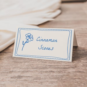 Pode incluir: Um cartão branco com uma borda azul e uma ilustração de uma flor azul. O texto "Cinnamon Scones" está escrito em azul cursivo.
