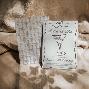 Op de afbeelding: Een witte uitnodigingskaart met een bruine illustratie van een martini-glas en de tekst "A tini bit older". De tekst "Olivia's 30th Birthday" staat onder de illustratie. De uitnodiging geeft de datum, tijd en locatie van het evenement aan.