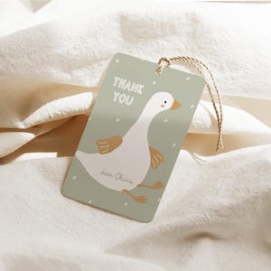 Pu&ograve; includere: Un'etichetta regalo rettangolare con un'illustrazione di un'oca dei cartoni animati. L'etichetta &egrave; verde chiaro con le parole "THANK YOU" in bianco. L'oca &egrave; bianca con dettagli marroni. Un cordino marrone &egrave; attaccato.