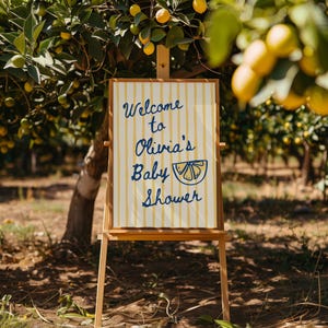 May include: A wooden easel displays a sign with the text "Welcome to Olivia's Baby Shower." The sign has a cream background with yellow vertical stripes and blue lettering. The scene is set outdoors with a lemon tree in the background.