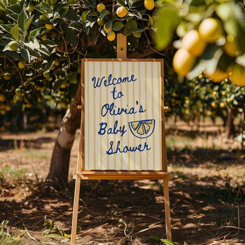 May include: A wooden easel displays a sign with the text "Welcome to Olivia's Baby Shower." The sign has a cream background with yellow vertical stripes and blue lettering. The scene is set outdoors with a lemon tree in the background.