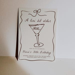 Op de afbeelding: Een witte uitnodigingskaart met bruine tekst en een tekening van een martini-glas. De tekst luidt "A tini bit older" en "Olivia's 30th Birthday". De uitnodiging beschrijft een feest op zaterdag 12 februari om 19:00 uur in The Rosy Ranch in Downtown, Boise. RSVP aan Lauren.