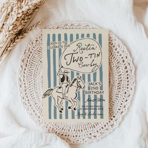 May include: A beige invitation card with blue vertical stripes and a cowboy illustration. The text reads "Rootin' Two-Tin Cowboy" and announces a 2nd birthday party. The card is on a white lace doily.