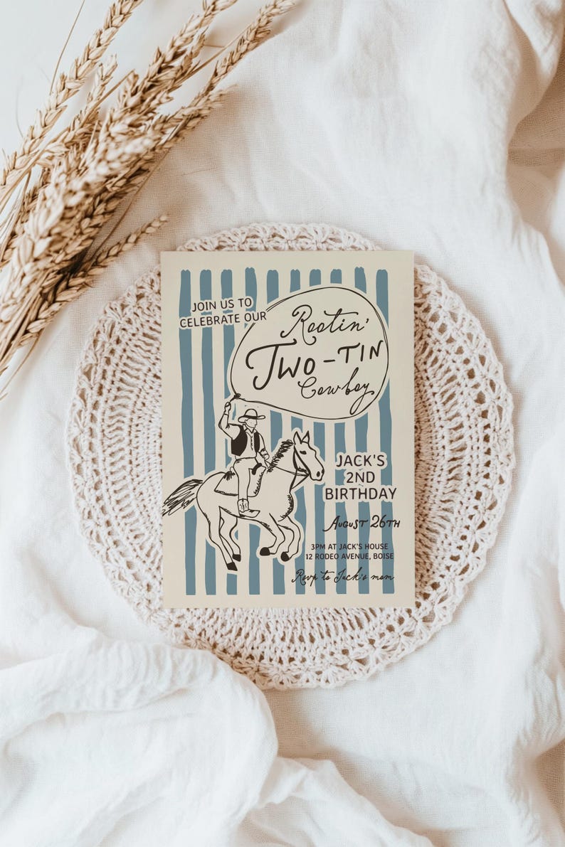 May include: A beige invitation card with blue vertical stripes and a cowboy illustration. The text reads "Rootin' Two-Tin Cowboy" and announces a 2nd birthday party. The card is on a white lace doily.