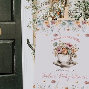 Może przedstawiać: Biały znak z kwiatowymi ilustracjami i tekstem "A baby is brewing - Welcome to Sadie's Baby Shower". Znak jest ustawiony na drewnianym sztalugach.