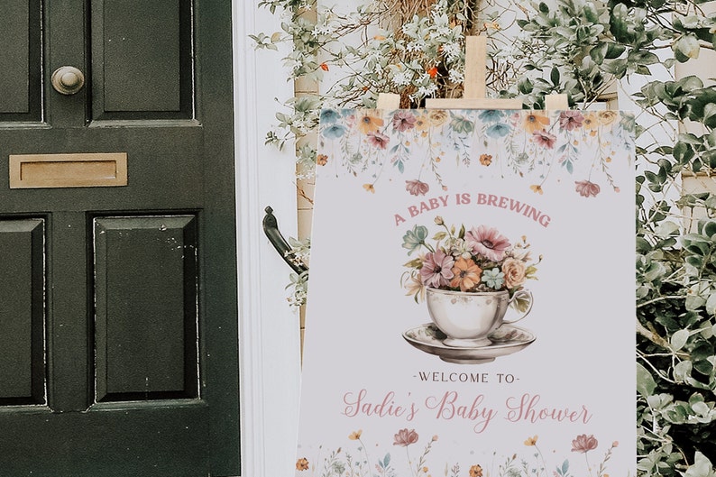 Może przedstawiać: Biały znak z kwiatowymi ilustracjami i tekstem "A baby is brewing - Welcome to Sadie's Baby Shower". Znak jest ustawiony na drewnianym sztalugach.
