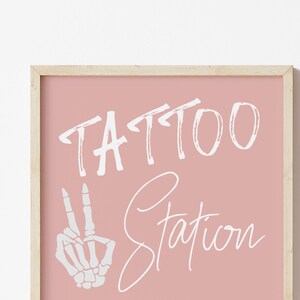 Puede incluir: Un cartel enmarcado en rosa con el texto "TATTOO STATION" en letras blancas. Una mano de esqueleto blanca con dos dedos levantados en se&ntilde;al de paz est&aacute; a la izquierda del texto.