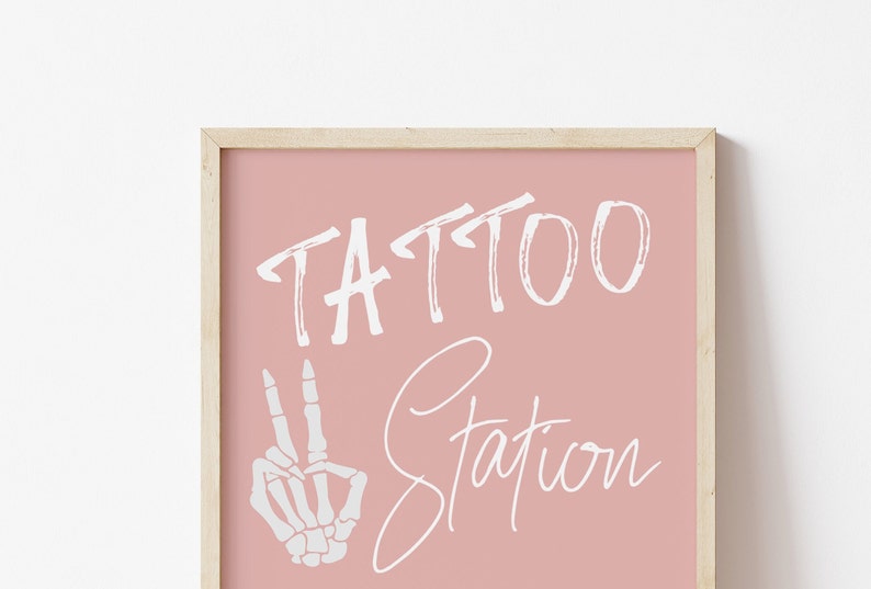Puede incluir: Un cartel enmarcado en rosa con el texto "TATTOO STATION" en letras blancas. Una mano de esqueleto blanca con dos dedos levantados en se&ntilde;al de paz est&aacute; a la izquierda del texto.
