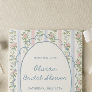 May include: A white card with a floral pattern and blue text that reads "Please join us for Olivia's Bridal Shower Saturday, July 10th".