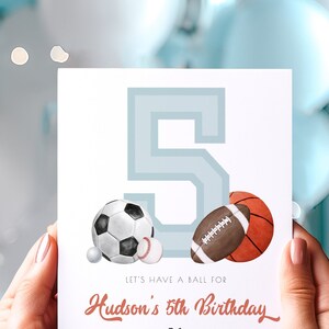 May include: A birthday card featuring a large number 5 in light blue, with illustrations of a soccer ball, baseball, football, and basketball. The text reads "Let's have a ball for Hudson's 5th Birthday."