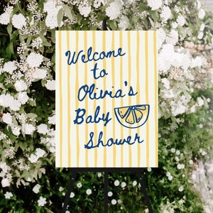 May include: A baby shower welcome sign on a black easel. The sign has a cream background with yellow vertical stripes. The text "Welcome to Olivia's Baby Shower" is in blue script, with a blue lemon slice illustration.