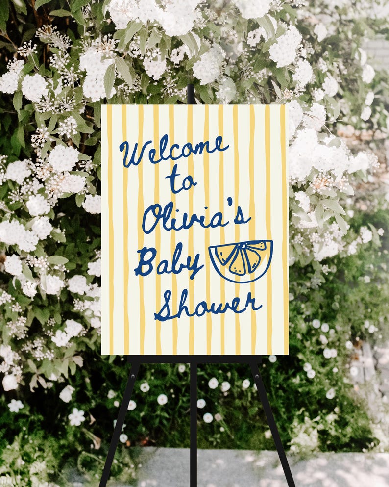 May include: A baby shower welcome sign on a black easel. The sign has a cream background with yellow vertical stripes. The text "Welcome to Olivia's Baby Shower" is in blue script, with a blue lemon slice illustration.