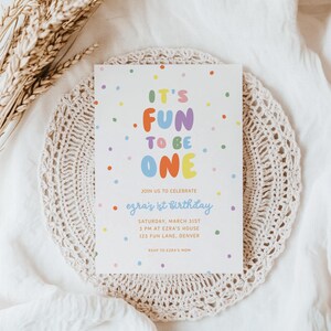 May include: A white invitation card with colorful text and polka dots, announcing "It's Fun to Be One" for Ezra's 1st birthday. The card is on a beige doily, with wheat stalks in the background.
