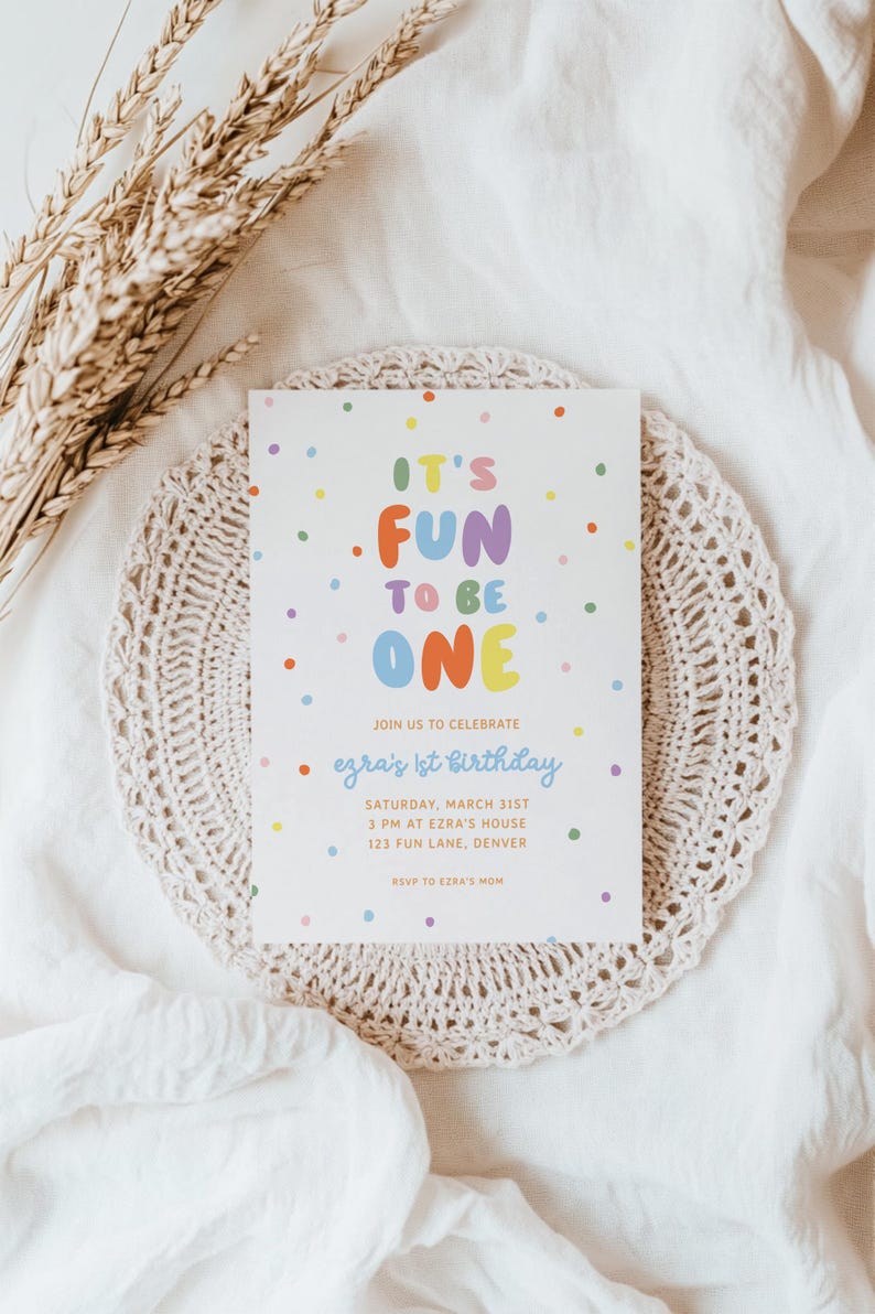 May include: A white invitation card with colorful text and polka dots, announcing "It's Fun to Be One" for Ezra's 1st birthday. The card is on a beige doily, with wheat stalks in the background.