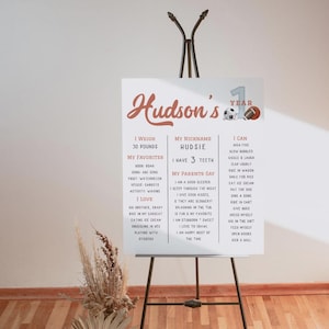 May include: A white poster with a red and white design celebrating a child's first birthday. The poster includes the child's name, Hudson, and details about their first year, including their weight, favorite activities, and things they can do.