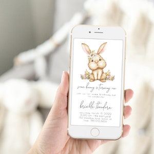 May include: A digital invitation for a first birthday party with a cute cartoon bunny wearing a flower crown. The invitation says "Some bunny is turning one" and includes the name "Brielle Theodora", the date "Saturday, March 19, 2022", the time "11:00 am", the location "The Barnyard, 153 Meadow Lane, Boise, ID", and the phone number "780.012.3456".