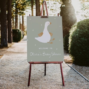 Pu&ograve; includere: Un cartello su un cavalletto di legno con la scritta "Welcome to Olivia's Baby Shower". Il cartello presenta un'oca dei cartoni animati con piume bianche, ali e zampe marroni e un becco giallo. Lo sfondo &egrave; verde chiaro.