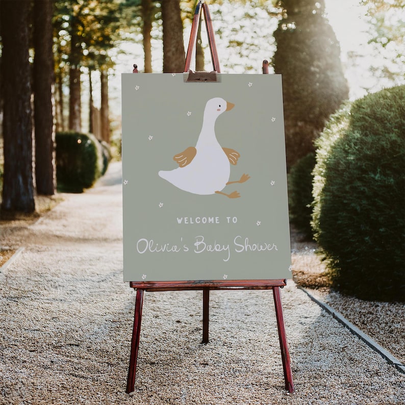 Pu&ograve; includere: Un cartello su un cavalletto di legno con la scritta "Welcome to Olivia's Baby Shower". Il cartello presenta un'oca dei cartoni animati con piume bianche, ali e zampe marroni e un becco giallo. Lo sfondo &egrave; verde chiaro.